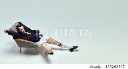 Fashion-forward young man stretching in chair with relaxed posture and vintage aesthetic against white studio background. Fashion-forward young man stretching in chair with relaxed posture and vintage aesthetic against white studio background. 123268327