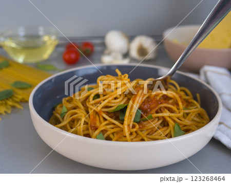 Lifting balanesi pasta with tomato sauce and basil from a modern bowl, surrounded by blurred ingredients like uncooked spaghetti and cherry tomatoes 123268464