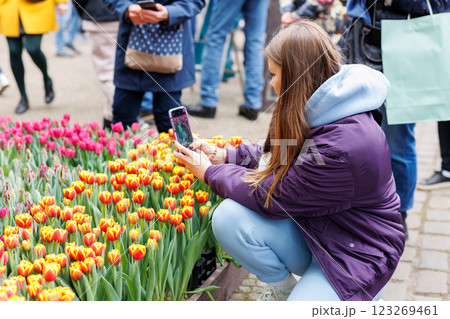 Young woman with smartphone leans over bright tulips in bustling outdoor event. She captures colorful blossoms while public gathers behind, enjoying vibrant spring display and seasonal beauty Young woman with smartphone leans over bright tulips in bustling outdoor event. She captures colorful blossoms while public gathers behind, enjoying vibrant spring display and seasonal beauty 123269461
