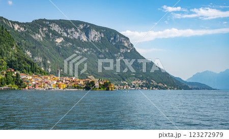 Menaggio, a charming town by Lake Como, showcases colorful buildings along the waterfront with lush green mountains providing a stunning backdrop under a bright blue sky. 123272979