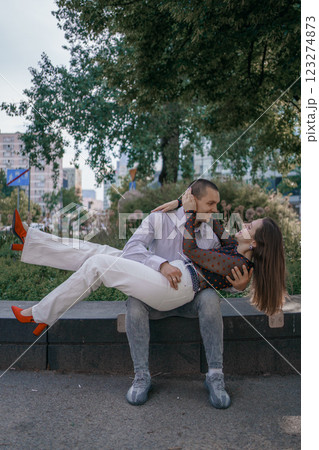 Happy couple in love enjoying a playful moment in the city park. A young man sitting on a bench holding his girlfriend in his arms as they smile at each other. Urban nature, romance, and joy. 123274873