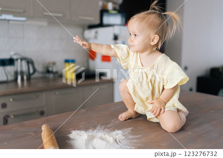 Playful toddler girl sitting amid scattered flour, reaching hands across messy kitchen table, exploring baking ingredients with curious expression. Concept of simple pleasures of childhood. 123276732