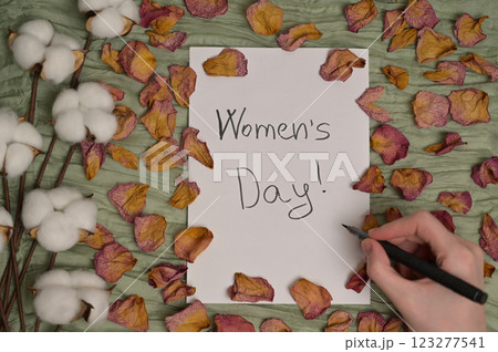 Woman's hand with a pen writing "Women's Day!" on white paper. Green cloth background with dry rose petals and cotton inflorescence plant. Greeting, invitation card. Top view 123277541