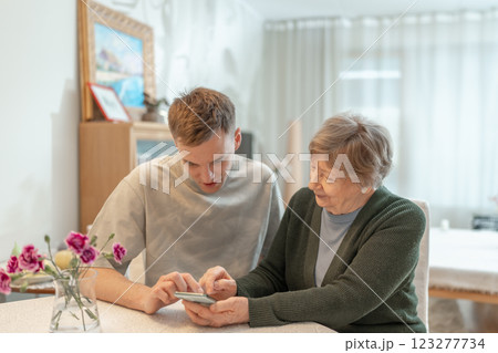 Young man helping a senior woman with a smartphone at home. Concept of technology, education, and senior digital adaptation. Grandson teaches grandmother how to use digital device. 123277734