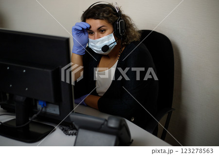 A call center agent working on the support hotline in the office, wearing a mask and gloves. The call center operator is talking to the customer. Close-up. 123278563
