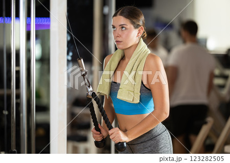 Girl in gym performs exercises for arm muscles using cable crossover machine and pull weighted rope Girl in gym performs exercises for arm muscles using cable crossover machine and pull weighted rope 123282505