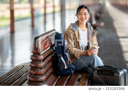 An Asian woman is smiling with the phone in hand while sitting on the wooden train station bench. 123285566