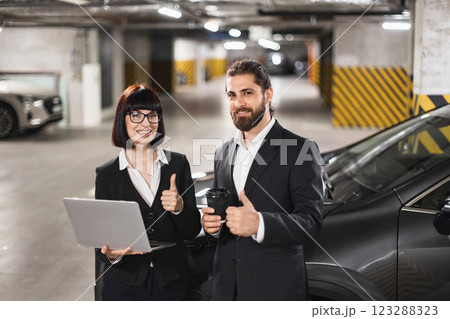 Caucasian businesswoman and businessman smiling, giving thumbs up in underground parking garage. Professional attire with laptop and coffee. Displays teamwork, success, confidence in urban office. Caucasian businesswoman and businessman smiling, giving thumbs up in underground parking garage. Professional attire with laptop and coffee. Displays teamwork, success, confidence in urban office. 123288323