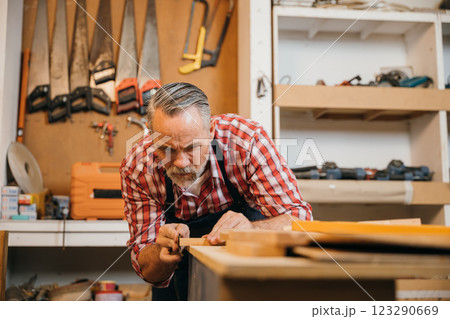 A senior carpenter in red plaid shirt carefully sands piece of wood in his workshop. Tools hang organized on wall behind him, illustrating his skill, dedication and attention to detail in woodworking A senior carpenter in red plaid shirt carefully sands piece of wood in his workshop. Tools hang organized on wall behind him, illustrating his skill, dedication and attention to detail in woodworking 123290669
