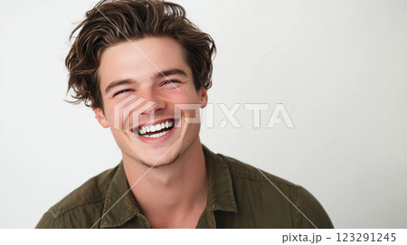 Cheerful young man with tousled brown hair smiles brightly, radiating positivity and joy against a simple white backdrop. Cheerful young man with tousled brown hair smiles brightly, radiating positivity and joy against a simple white backdrop. 123291245