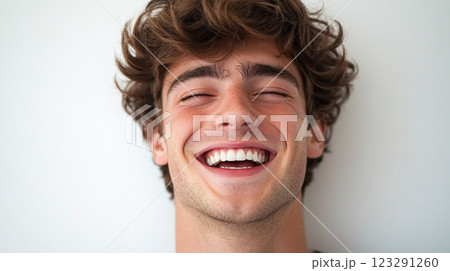 A young man with curly hair is laughing joyously, his eyes closed and face beaming with happiness in front of a plain backdrop. 123291260
