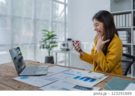 Happy young woman with smartphone at desk celebrating success in a modern office workspace filled with documents and plants 123291889