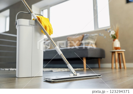Cleaning supplies: mop, gloves, bucket with wringer sitting on wooden laminate floor in modern living room, wet equipment tools, housekeeping concept. Emphasizing home sanitation, cleanliness, hygiene Cleaning supplies: mop, gloves, bucket with wringer sitting on wooden laminate floor in modern living room, wet equipment tools, housekeeping concept. Emphasizing home sanitation, cleanliness, hygiene 123294371