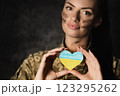 Military woman in military uniform with camouflage makeup on her face holds a heart-shaped Ukrainian flag in hands. Close-up 123295262