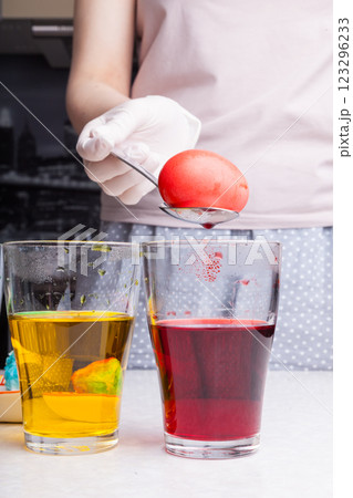 Preparing for Easter - coloring eggs in home kitchen female hands in rubber gloves use spoon take boiled egg out of glass with diluted red dye next other glasses different colors with submerged eggs. 123296233