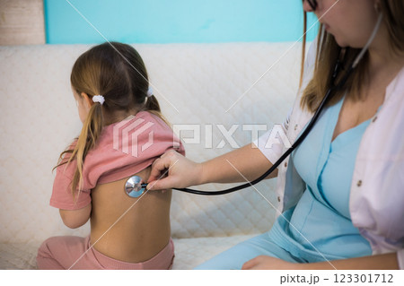 A pediatrician visits a sick child at home. A female pediatrician uses a stethoscope to examine the lungs of a sick little girl as she sits on a couch at home during a medical home visit. 123301712