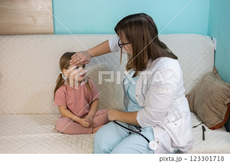 A sick child has a doctor called to her home. A female pediatrician visits the little girl at home and measures her temperature by placing her hand on her forehead while she sits on the couch. A sick child has a doctor called to her home. A female pediatrician visits the little girl at home and measures her temperature by placing her hand on her forehead while she sits on the couch. 123301718