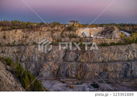 A rocky cliff with sparse trees and dry bushes under warm sunlight. At the top, an abandoned industrial structure stands among autumn foliage, creating a contrast between nature and human-made ruins. 123301889