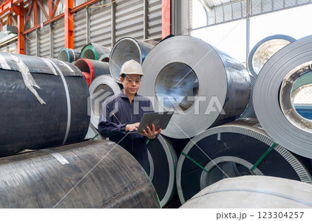 A man in a hardhat and mechanic coverall dust protection suit checks laptop computer within the metal sheet factory. Steel sheet roll stack are in the working area. A man in a hardhat and mechanic coverall dust protection suit checks laptop computer within the metal sheet factory. Steel sheet roll stack are in the working area. 123304257