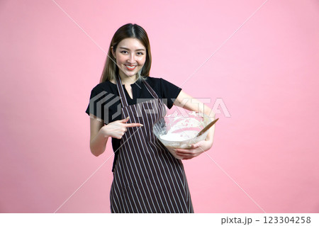 A happy woman in an apron, pointing finger at batter in a clear glass bowl with a wooden spoon. Portrait on pink background with studio light. 123304258