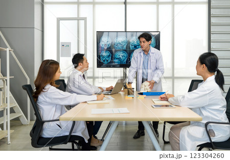 Young medical professional standing and presenting, holding a model of human skull and referencing image of brain scan displayed on a screen. The audience listening attentively with the presentation. Young medical professional standing and presenting, holding a model of human skull and referencing image of brain scan displayed on a screen. The audience listening attentively with the presentation. 123304260