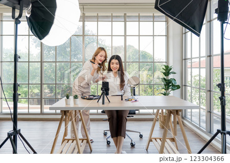 Two asian women creating content for social media. They are using a studio setup that include a smartphone mounted on a tripod, lighting equipment and various makeup product arrayed on the table. 123304366