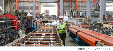Two workers collaborate in a busy factory, examining machinery with metal coils in the background. The environment is well-lit and organized, showcasing industrial activity. 123304485