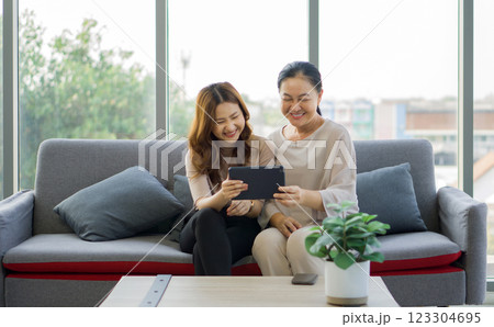 Two women are sitting on a couch, laughing together and sharing a tablet computer, in a bright living room. Two women are sitting on a couch, laughing together and sharing a tablet computer, in a bright living room. 123304695