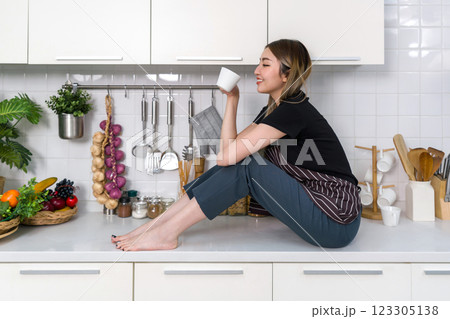 Young asian woman with a striped apron drink coffee from mug while sitting on counter in a modern kitchen. Cooking utensil and an assortment of decorated fruit and vegetable are on the countertop. Young asian woman with a striped apron drink coffee from mug while sitting on counter in a modern kitchen. Cooking utensil and an assortment of decorated fruit and vegetable are on the countertop. 123305138