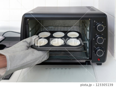 Hand with oven gloves, placing a muffin tray with batter into a black toaster oven in a white kitchen. 123305139