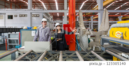 Two worker with safety helmet and black suit standing in front of metal sheet roofing machine, holding tablet computer used for taking note, checking schedule and reviewing digital document. Two worker with safety helmet and black suit standing in front of metal sheet roofing machine, holding tablet computer used for taking note, checking schedule and reviewing digital document. 123305140