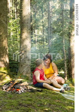 A woman and child sit by a campfire in a forest, sharing a quiet moment amidst the trees. 123308304