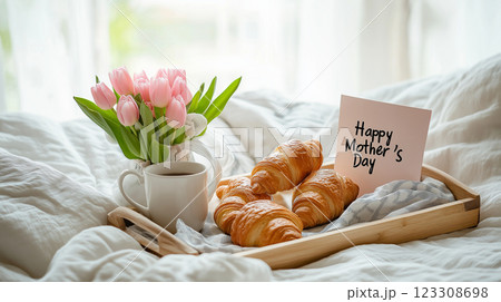 A cozy Mothers Day breakfast in bed with fresh croissants, coffee, pink tulips, and a handwritten card on a wooden tray. 123308698