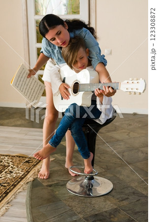 A mother helps her child learn to play the guitar, showing them sheet music and guiding their hands. A mother helps her child learn to play the guitar, showing them sheet music and guiding their hands. 123308872