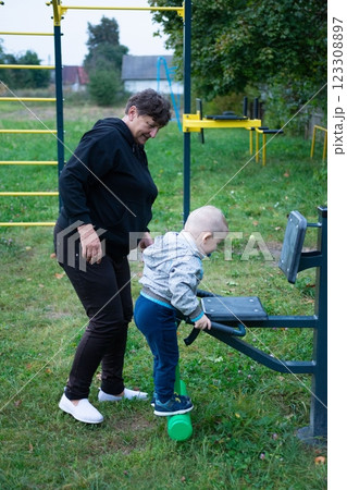 Grandmother assists toddler at outdoor gym, showing encouragement and support as the child explores equipment on a grassy field. Grandmother assists toddler at outdoor gym, showing encouragement and support as the child explores equipment on a grassy field. 123308897