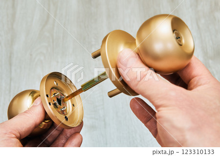 Door knob assembly, partly disassembled, reveals square spindle connecting two brass knobs, showcasing internal mechanism and human hand holding parts. 123310133