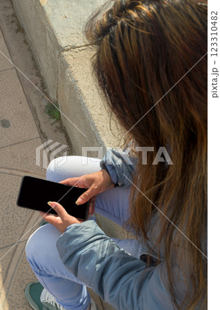 latina woman sitting on a stone bench, looking at a smartphone, dressed casually, mallorca balearic islands, spain 123310482