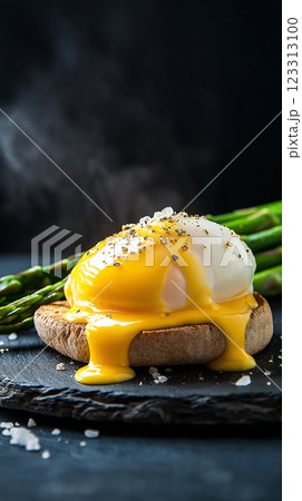 indulgent eggs benedict with poached egg and golden hollandaise sauce on rustic stone plate, fresh asparagus spears, dark slate background, gourmet breakfast, moody food photography 123313100