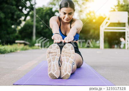 Sporty girl stretching her legs sitting on a mat in a park. Athlete woman sitting doing leg stretch in a park. Athlete girl sitting on a mat stretching her legs in a park 123317328