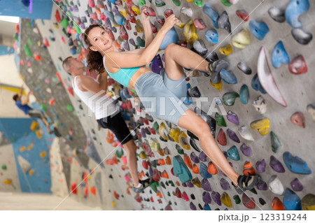 Adult woman is mastering climbing on training wall in gym, side view. Young woman holds on tightly to ledges and strives for top of bouldering route 123319244