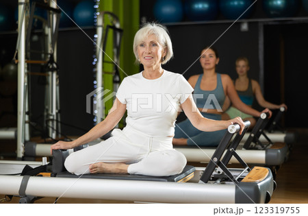 Mature woman sitting on foldable bed reformer performs exercises, strengthening body and muscles Mature woman sitting on foldable bed reformer performs exercises, strengthening body and muscles 123319755