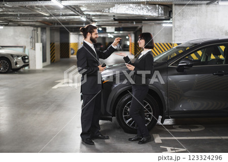 Caucasian businessman selling car to Caucasian woman in underground parking lot. Man and woman, both in business attire, exchanging car keys with evident professionalism and agreement. Caucasian businessman selling car to Caucasian woman in underground parking lot. Man and woman, both in business attire, exchanging car keys with evident professionalism and agreement. 123324296