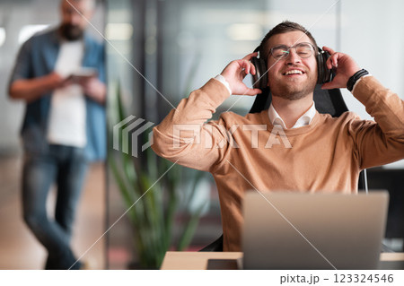 Excited businessman wearing headphones, sitting at his desk in a modern open-space startup office. Overjoyed executive enjoying his break, singing and listening to favorite music, having fun at work. Excited businessman wearing headphones, sitting at his desk in a modern open-space startup office. Overjoyed executive enjoying his break, singing and listening to favorite music, having fun at work. 123324546
