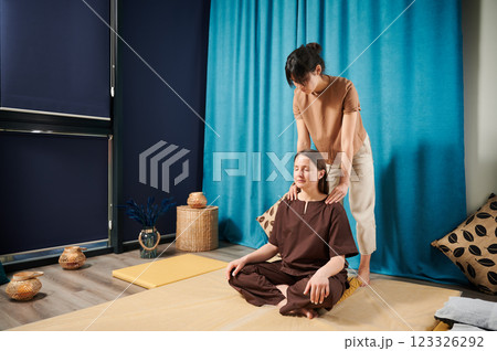 Masseuse making thai yoga massage. Woman therapist doing traditional massage treatment for female patient indoors. Client sitting on mat, meditating, enjoying hand massage in calm atmosphere. Masseuse making thai yoga massage. Woman therapist doing traditional massage treatment for female patient indoors. Client sitting on mat, meditating, enjoying hand massage in calm atmosphere. 123326292
