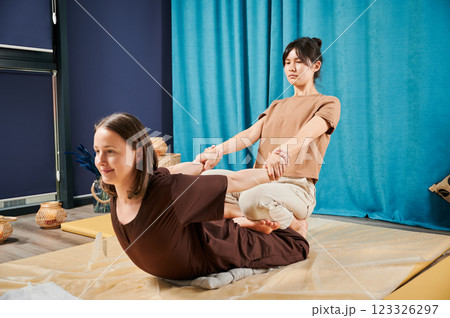 Masseuse making thai yoga massage. Woman therapist doing traditional massage treatment, stretching female patient's body. Client lying on mat, enjoying hand massage in calm atmosphere. Masseuse making thai yoga massage. Woman therapist doing traditional massage treatment, stretching female patient's body. Client lying on mat, enjoying hand massage in calm atmosphere. 123326297