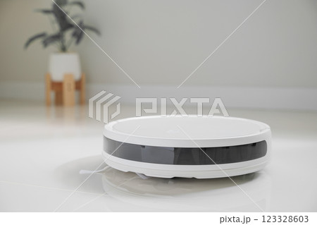 White automatic floor cleaner with side brush on tile flooring, featuring front black sensor strip. Robot vacuum cleaner efficiently cleaning floor, illustrating smart home technology for dust removal White automatic floor cleaner with side brush on tile flooring, featuring front black sensor strip. Robot vacuum cleaner efficiently cleaning floor, illustrating smart home technology for dust removal 123328603