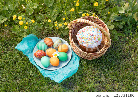 Easter cake in a wicker basket and a plate with colored Easter eggs on green grass with yellow Persian buttercup. Easter cake in a wicker basket and a plate with colored Easter eggs on green grass with yellow Persian buttercup. 123331190