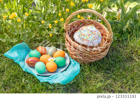 Easter cake in a wicker basket and a plate with colored Easter eggs on green grass with yellow Persian buttercup. Easter cake in a wicker basket and a plate with colored Easter eggs on green grass with yellow Persian buttercup. 123331191