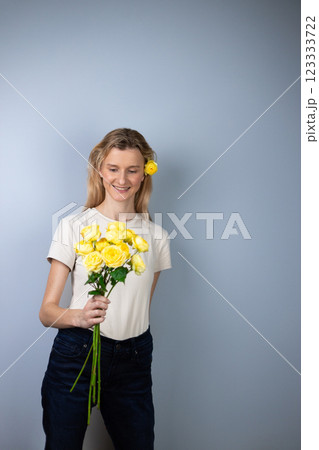 on a light gray background a young blonde girl in a light T-shirt in her hands yellow peony roses March 8 smile on her face bud in her hair looking at the camera 123333722