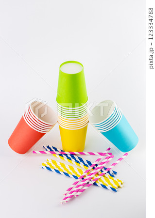 white background, a stack of paper cups for coffee and tea painted in red and blue rest on yellow and green disposable tableware for recreation and picnics eco cups striped cocktail straws 123334788